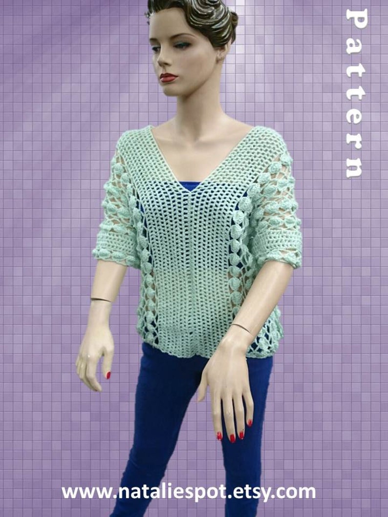 Peut inclure: Un haut en crochet vert clair avec un col en V et des manches courtes. Le haut pr&eacute;sente un motif ajour&eacute; d&eacute;licat et de petites fleurs en crochet autour du col et des manches. Le haut est port&eacute; par un mannequin sur un fond violet.
