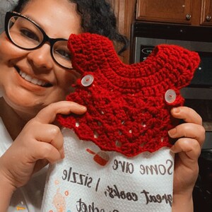 May include: A white kitchen towel with a red crocheted dishcloth on top. The towel has a design of yarn balls and a cat holding a ball of yarn. The text on the towel reads "Hook! With a crochet, great cooks, some give size!"