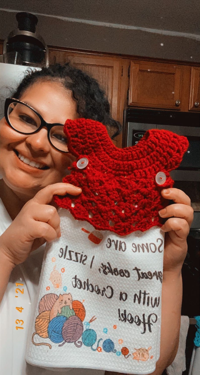 May include: A white kitchen towel with a red crocheted dishcloth on top. The towel has a design of yarn balls and a cat holding a ball of yarn. The text on the towel reads "Hook! With a crochet, great cooks, some give size!"