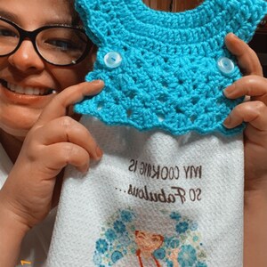 May include: White kitchen towel with a blue floral design and the text "Cheers to me! Even the smoke the air!"  A woman in a red apron is featured in the center of the design. The towel has a blue crocheted potholder attached to the top.
