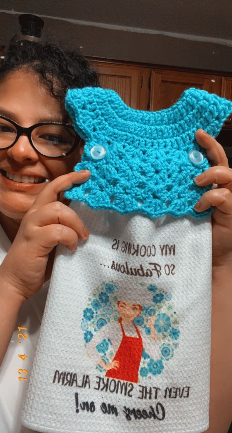 May include: White kitchen towel with a blue floral design and the text "Cheers to me! Even the smoke the air!"  A woman in a red apron is featured in the center of the design. The towel has a blue crocheted potholder attached to the top.