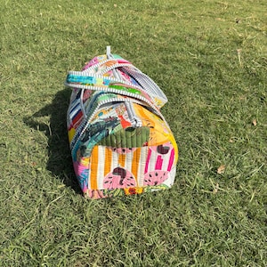 May include: A vibrant, quilted duffel bag with a tropical design and striped details. The bag displays a mix of colours, including pink, yellow, green, and blue. The bag is placed on a patch of green grass.
