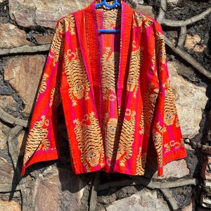 Op de afbeelding: Een levendige, open kimono-achtige jas in tinten rood, roze en oranje, versierd met een herhalend tijgerpatroon. De jas is gemaakt van een lichte stof en heeft een contrasterende roze rand. Hij hangt aan een blauwe hanger.