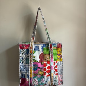 May include: A quilted tote bag featuring a vibrant patchwork design with various floral, strawberry, and striped patterns in red, blue, green, and yellow. The bag has two long, sturdy handles with a black and white striped trim along the edges.
