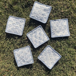 May include: Six square quilted coasters with a white background and blue floral pattern. Each coaster has a blue and white striped border. The coasters are arranged on a bed of green grass.