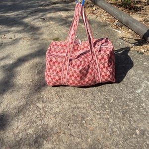 Könnte beinhalten: Eine rosa Tragetasche mit floralem Muster und passenden Griffen und Besatz. Die Tasche hat eine rechteckige Form mit einer Tasche auf der Vorderseite. Die Tasche ist aus einem gesteppten Stoff gefertigt und hängt an einem blauen Haken.