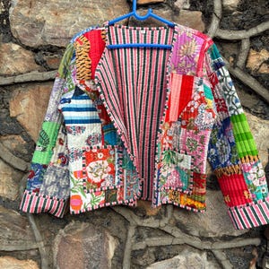 Può includere: Giacca patchwork con una varietà di fantasie colorate, tra cui floreali, a righe e animalier. La giacca aperta è appesa a una gruccia blu. Le maniche e i bordi sono rifiniti con tessuto a righe.