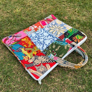 Op de afbeelding: Een kleurrijke patchwork laptoptas met een witte rits en een handvat met patroon. De tas heeft verschillende stofpatronen in rood, blauw, groen en roze, tegen een achtergrond van groen gras.
