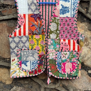 May include: A patchwork vest with a variety of floral and geometric patterns in shades of pink, green, orange, and blue. The vest has a striped lining and trim, and two front pockets. It is hanging on a blue hanger.
