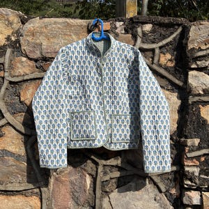 May include: A quilted jacket with a white background and a blue floral pattern. The jacket has a mandarin collar, two front pockets, and green trim. It is hanging on a blue hanger against a stone wall.