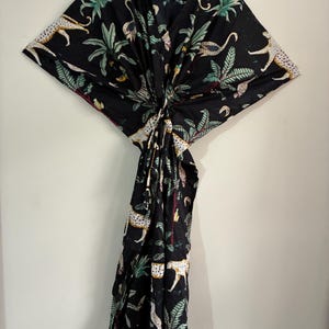 May include: A black kaftan dress featuring a repeating pattern of white and yellow cheetahs interspersed with green palm leaves and tropical foliage. The garment has a V-neckline and a central knotted detail at the waist, creating a draped silhouette.