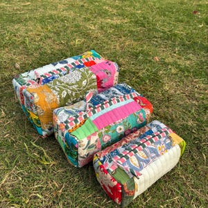 以下が含まれることがあります： 緑の草の上に、花柄とパッチワークのデザインが施された、カラフルな長方形のキルトバッグが3つあります。 バッグはピンク、緑、青、黄色、赤など、さまざまな色を使用しています。 バッグは手作りのようです。