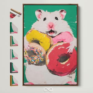 Op de afbeelding: Een ingelijste canvas print met een witte hamster met een roze neus en open mond, die kleurrijke donuts vasthoudt. De donuts zijn geel, roze, rood en een met hagelslag, tegen een groene achtergrond.
