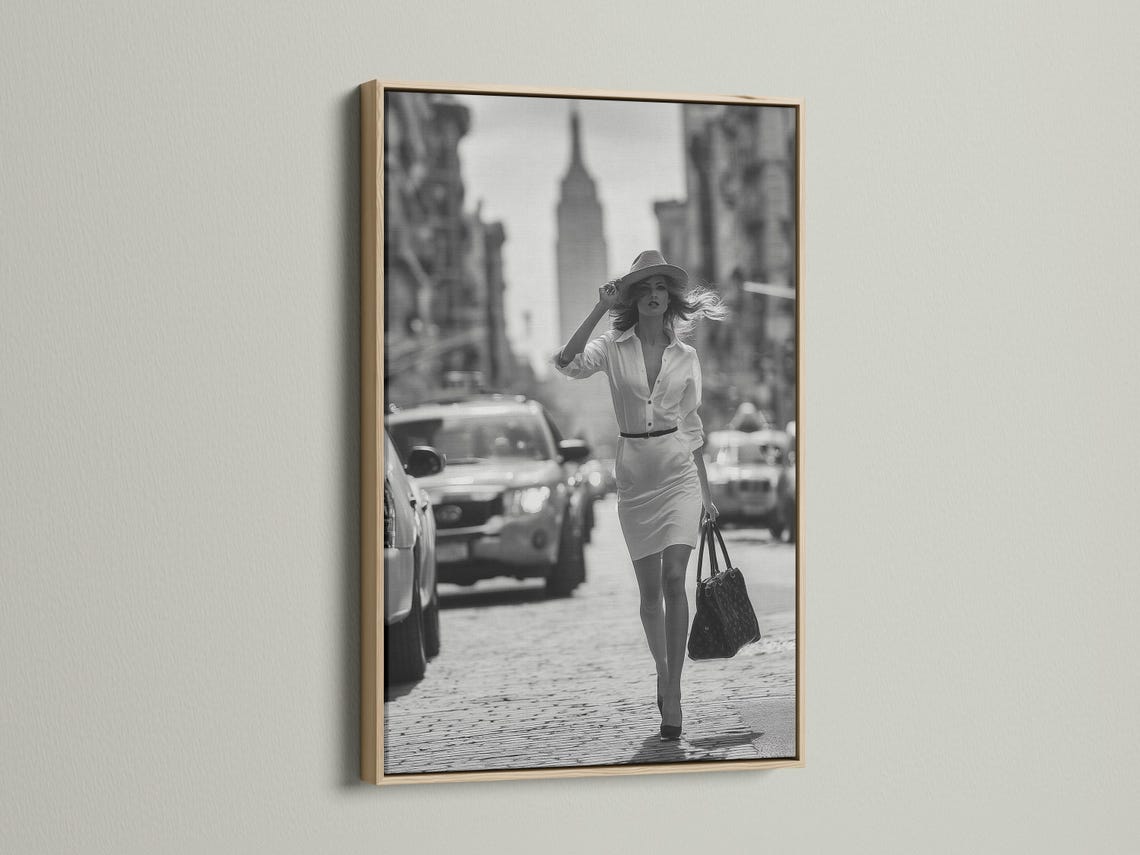 A black-and-white street photograph shows a stylish woman walking forward with a hat and handbag, with blurred city buildings and cars behind her
