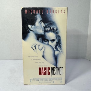 May include: A VHS tape of the movie "Basic Instinct" featuring Michael Douglas and Sharon Stone. The cover art shows a man and woman in a close embrace, with the film's title in red. The tape is in a white case.