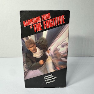 May include: A VHS tape box for the movie "The Fugitive" starring Harrison Ford. The box is black with red and white text. The image on the box shows Harrison Ford running. The text on the box reads "A murdered wife. A one-armed man. An obsessed detective. The chase begins."