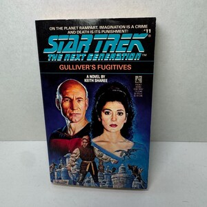 Puede incluir: Un libro de bolsillo titulado "Star Trek: The Next Generation: Gulliver's Fugitives" de Keith Sharee. La portada presenta retratos de dos personas y una figura con un atuendo marrón. El libro tiene un fondo azul y negro con el texto "¡En el planeta Rampart, la imaginación es un crimen y la muerte es su castigo!"