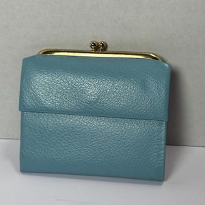 May include: A teal-colored wallet with a kiss-lock clasp. The wallet has a textured surface and a gold-colored frame. The clasp is a small, round design. The wallet is closed and appears to be made of leather.