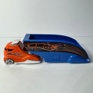 May include: A die-cast toy truck featuring an orange cab with blue flame accents and a detachable blue trailer. The trailer side displays black and orange flame graphics with the Hot Wheels logo printed in white and orange lettering.