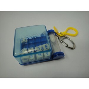 May include: A miniature Boggle game keychain featuring a translucent blue plastic cube case containing small white lettered dice. Attached to the side is a small blue hourglass timer and a yellow plastic clip with a silver metal carabiner.