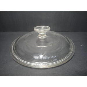 Puede incluir: Una tapa de vidrio circular y transparente para utensilios de cocina con un pomo central. El borde de la tapa tiene grabado el texto CORNING NY USA. El vidrio es transparente y presenta una superficie lisa y reflectante.