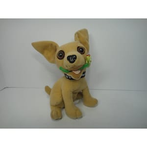 Puede incluir: Un peluche de perro chihuahua color canela con grandes ojos marrones, orejas erguidas y un collar negro con texto en blanco. El perro de peluche sostiene una pequeña rosa de tela verde y roja en la boca, con una lengua color canela visible.