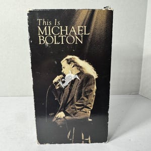 May include: A vintage VHS tape case with the title "This Is MICHAEL BOLTON" in white text. The image features a sepia-toned photo of a man with long, wavy hair holding a microphone. The case is black with some wear.