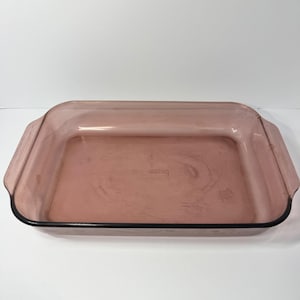 May include: A rectangular baking dish made of translucent pink glass with integrated handles on both short ends. The center of the dish features embossed text that reads Pyrex, indicating the brand and the specific glass material used.