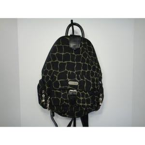 May include: A black and olive green giraffe-patterned backpack featuring a top handle, a front pocket with a silver-toned buckle closure, two side zippered pockets, and silver-toned hardware accents. The bag has a structured shape and adjustable straps.