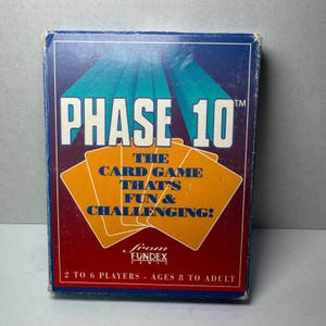 Könnte beinhalten: Eine Phase 10 Kartenspielbox mit blauem und rotem Hintergrund. Der Spieltitel "PHASE 10" steht in großen weißen Buchstaben. Die Box enthält auch den Text "THE CARD GAME THAT'S FUN & CHALLENGING!" und "2 TO 6 PLAYERS - AGES 8 TO ADULT."