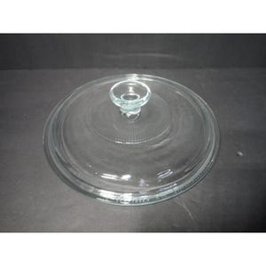 Puede incluir: Una tapa de cristal circular y transparente con un pomo central. La tapa presenta una superficie lisa con un sutil patrón de estrías radiantes alrededor de la base del pomo y un borde elevado a lo largo del contorno exterior.
