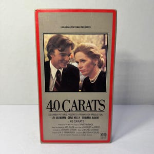 以下が含まれることがあります： 映画「40 Carats」のヴィンテージVHSテープ。箱は長方形で、赤い縁と灰色の背景があります。カバーには2人の写真と映画のタイトルが写っています。「Columbia Pictures Presents」の文字が上部にあります。