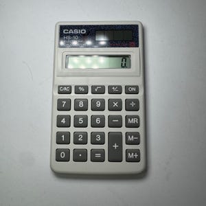 May include: A Casio HS-10 electronic calculator with a light gray body and dark gray buttons. The calculator has a digital display showing "0." The keys include numbers, basic math functions, and memory functions.