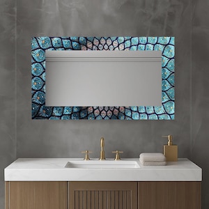 May include: Rectangular mirror with a decorative blue and teal floral border. The mirror is above a white countertop with gold taps and a soap dispenser. The bathroom features a wooden cabinet and a neutral colour scheme.