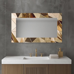 Op de afbeelding: Rechthoekige spiegel met een geometrisch gouden frame. Het frame heeft een mozaïek van driehoekige vormen in goud- en bronstinten. De spiegel is gemonteerd boven een wit aanrecht met gouden armaturen en een houten kast.