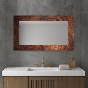 Puede incluir: Un espejo rectangular con un marco de vetas de madera natural cuelga sobre una encimera blanca. El espejo refleja una pared gris y una moldura blanca. Un grifo dorado, un dispensador de jabón y una toalla doblada están sobre la encimera.