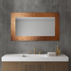 Pode incluir: Um espelho retangular cor de cobre com uma superfície texturizada está pendurado acima de uma bancada branca. A moldura do espelho tem uma aparência áspera e martelada. Abaixo, uma torneira dourada e um dispensador de sabão estão na bancada, ao lado de uma toalha dobrada.