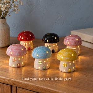 May include: Six ceramic mushroom-shaped lamps in red, black, pink, blue, and yellow, each featuring white spots and illuminated windows. The lamps are arranged on a wooden surface with the text "Find your favourite little glow" at the bottom.