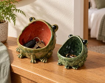 Può includere: Due ciotole in ceramica verde a forma di rana con pelle testurizzata e occhi neri. La ciotola più grande ha un interno rosso-arancio contenente un mazzo di chiavi in metallo. La più piccola ha un interno verde scuro con un anello dorato.