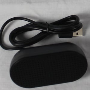 Può includere: Un altoparlante USB nero con una superficie strutturata e un cavo USB. L'altoparlante è di forma ovale. Nella foto è presente anche un sacchetto di plastica trasparente con l'etichetta "USB Speaker".