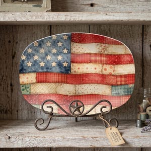 May include: Oval decorative plate featuring a distressed American flag design. The plate has a blue field with white stars and red and white stripes. It is displayed on a wrought iron stand with a star accent. The tag reads "Primitive Americana Collection."