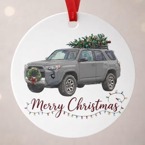 May include: Round Christmas ornament featuring a gray SUV with a Christmas tree on top and a wreath on the front. The words "Merry Christmas" are written in cursive below the car, with a string of colorful lights. A red ribbon is at the top.