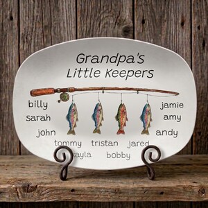 Peut inclure: Une assiette décorative ovale blanche avec le texte "Grandpa's Little Keepers" et des noms sous une canne à pêche avec des poissons suspendus. Les noms incluent Billy, Sarah, John, Tommy, Kayla, Tristan, Jared, Bobby, Jamie, Amy et Andy.