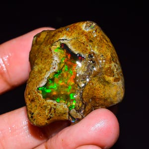 Könnte beinhalten: Ein rohes, unpoliertes Boulder-Opal-Exemplar mit einer rauen, texturierten, beigefarbenen Außenseite und einer zentralen Aushöhlung, die lebhafte, schillernde Blitze in Neongrün und Feuerrot vor einer dunklen Matrix zeigt.