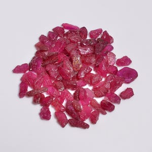 Könnte beinhalten: Ein Haufen roher, ungeschliffener Rubin-Edelsteine in verschiedenen Rot- und Rosatönen. Die Edelsteine haben eine raue, natürliche Textur und sind auf einer weißen Oberfläche verstreut. Die Edelsteine sind in verschiedenen Größen und Formen.