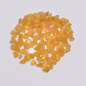 Könnte beinhalten: Eine Ansammlung kleiner, roher und unpolierter gelber und orangefarbener Edelsteine. Diese losen Steine haben unregelmäßige, gezackte Formen mit einem durchscheinenden bis halb-durchscheinenden Aussehen und zeigen verschiedene Schattierungen von Goldgelb, Bernstein und hellem Orange.