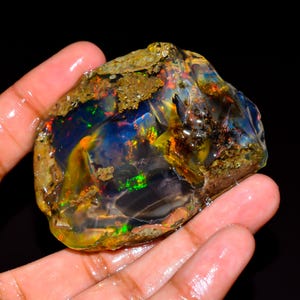 May include: A raw, unpolished boulder opal specimen featuring a dark, translucent base with vibrant flashes of iridescent green, orange, and red play-of-colour. The stone has an irregular, organic shape with rough, earthy matrix textures along the edges.
