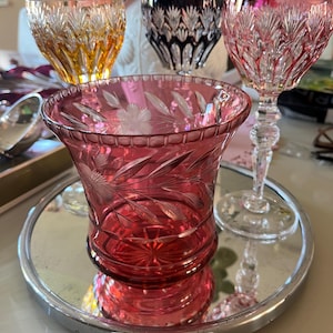 Op de afbeelding: Een verzameling sierlijke drinkglazen van geslepen glas in verschillende kleuren. Een robijnrode vaas met geëtste bloemmotieven staat op een spiegelende tray. Andere glazen zijn in amber-, zwarte en roze tinten, met ingewikkelde patronen.