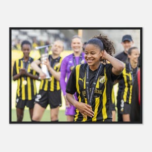 May include: A footballer wearing a black and yellow striped shirt with a medal around their neck. The shirt features a team crest and text. In the background, other players in similar kits are visible, one holding a silver trophy.