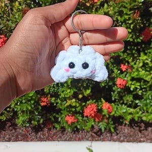 Puede incluir: Un llavero blanco tejido en forma de nube con ojos negros y mejillas rosas. El llavero está sujeto a un anillo de metal plateado. El fondo presenta follaje verde y flores rojas.